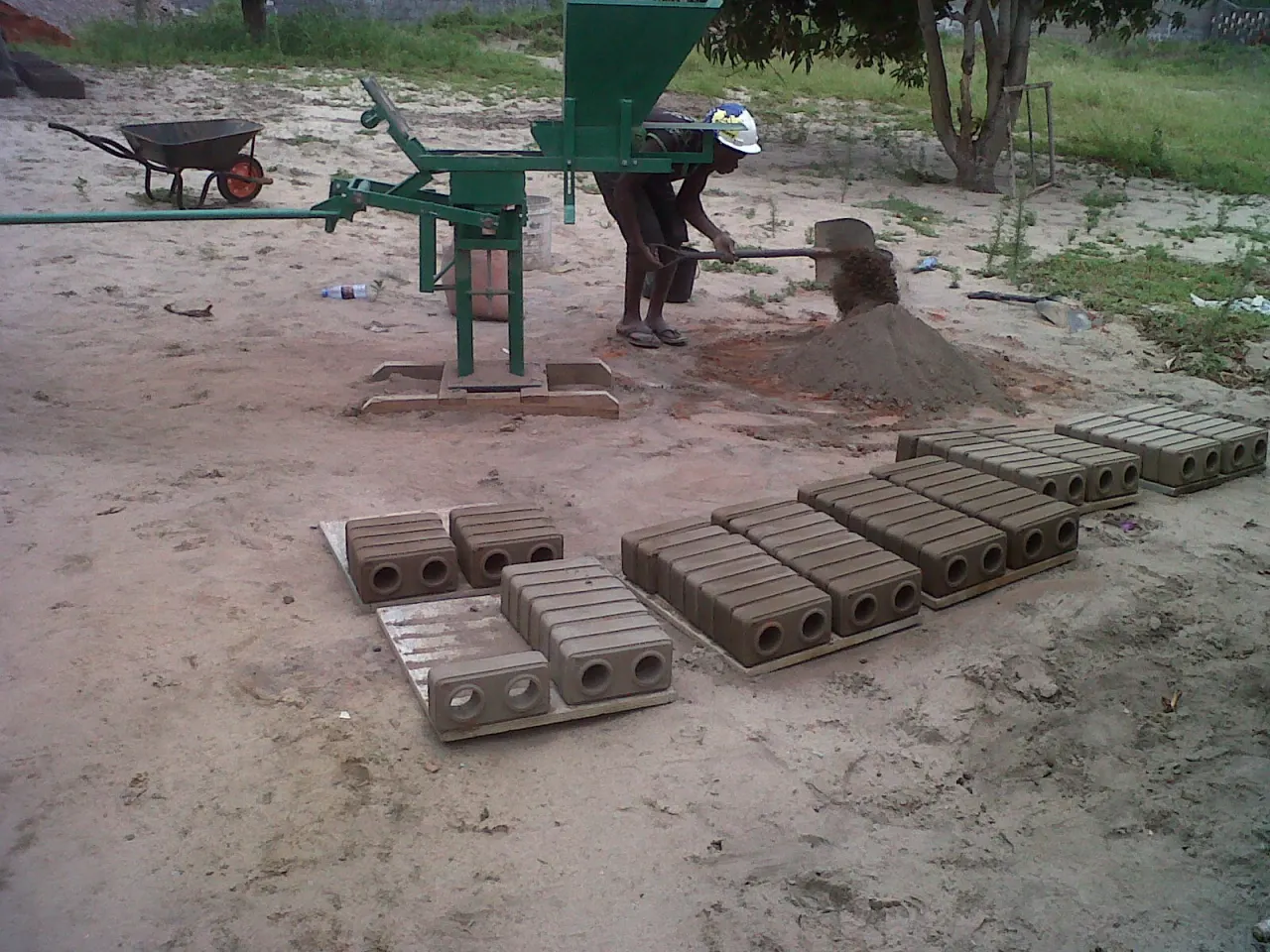 A worker shoveling soil next to a press, with freshly pressed ecological bricks on the ground